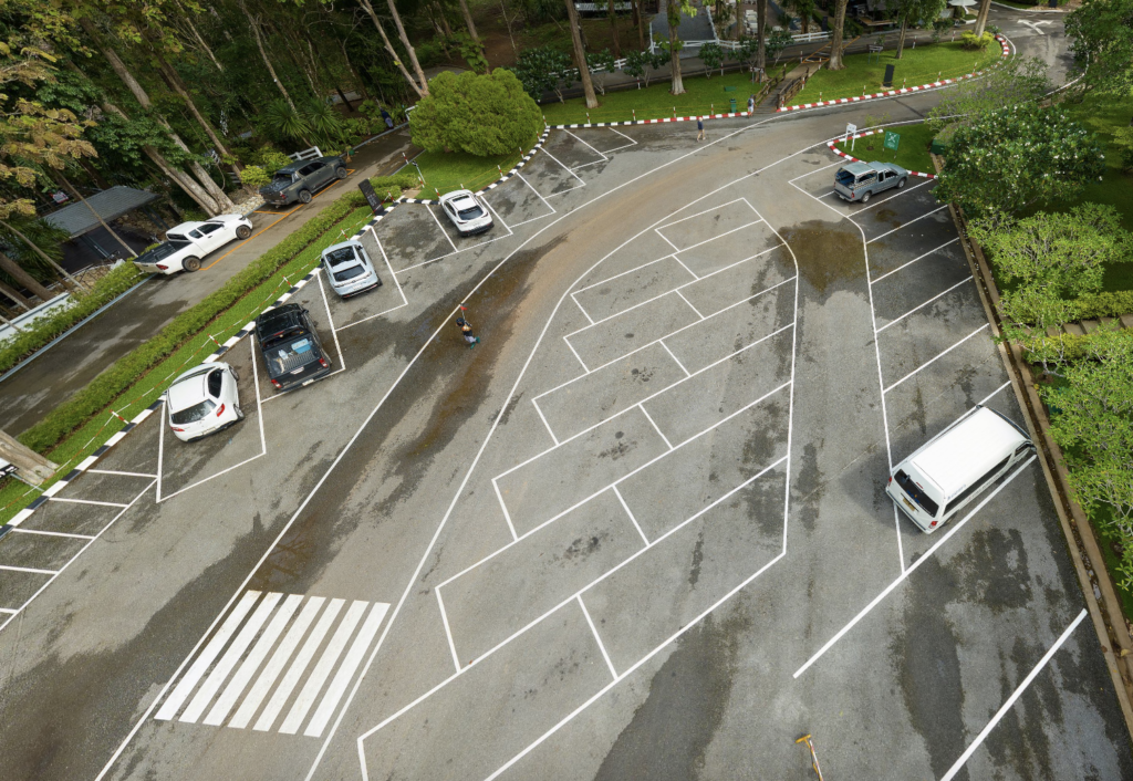 8 cars on grey ground with white markings surrounded by green vegetation and trees.