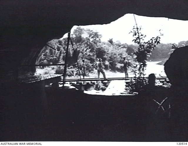 A group of people in a very large cave looking out at a soldier on the railway line bridge.