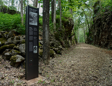 Information sign at entrance to rocky pathway surrounded by rock walls and green vegetation.
